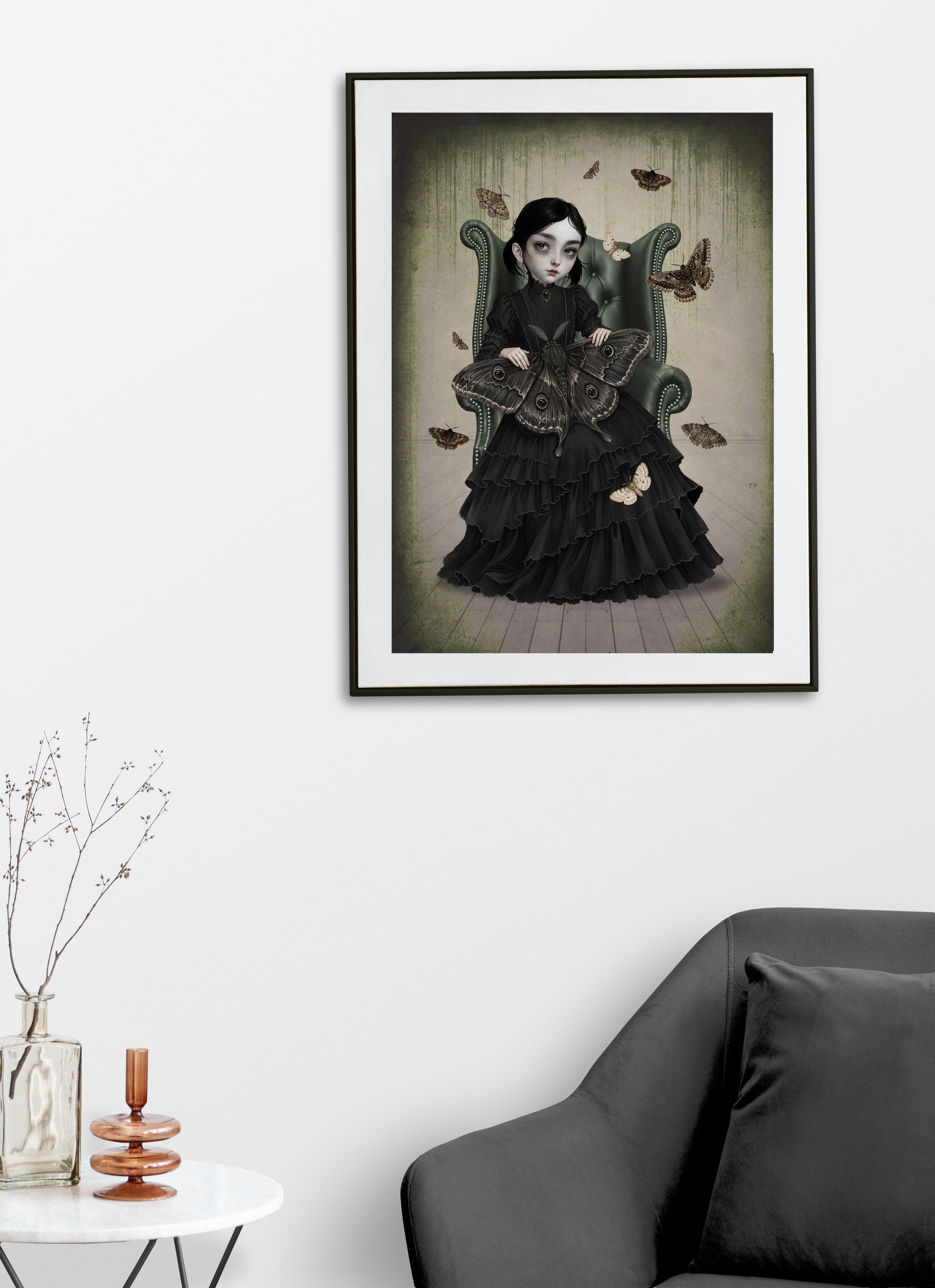 A framed art print featuring a gothic girl in a black dress sitting in a green armchair with moths displayed on a white wall above a dark gray sofa and a small side table with decorative items.