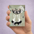 Hand holding an ACEO art card with a design of a twin werewolf sisters and a full moon on a light purple background