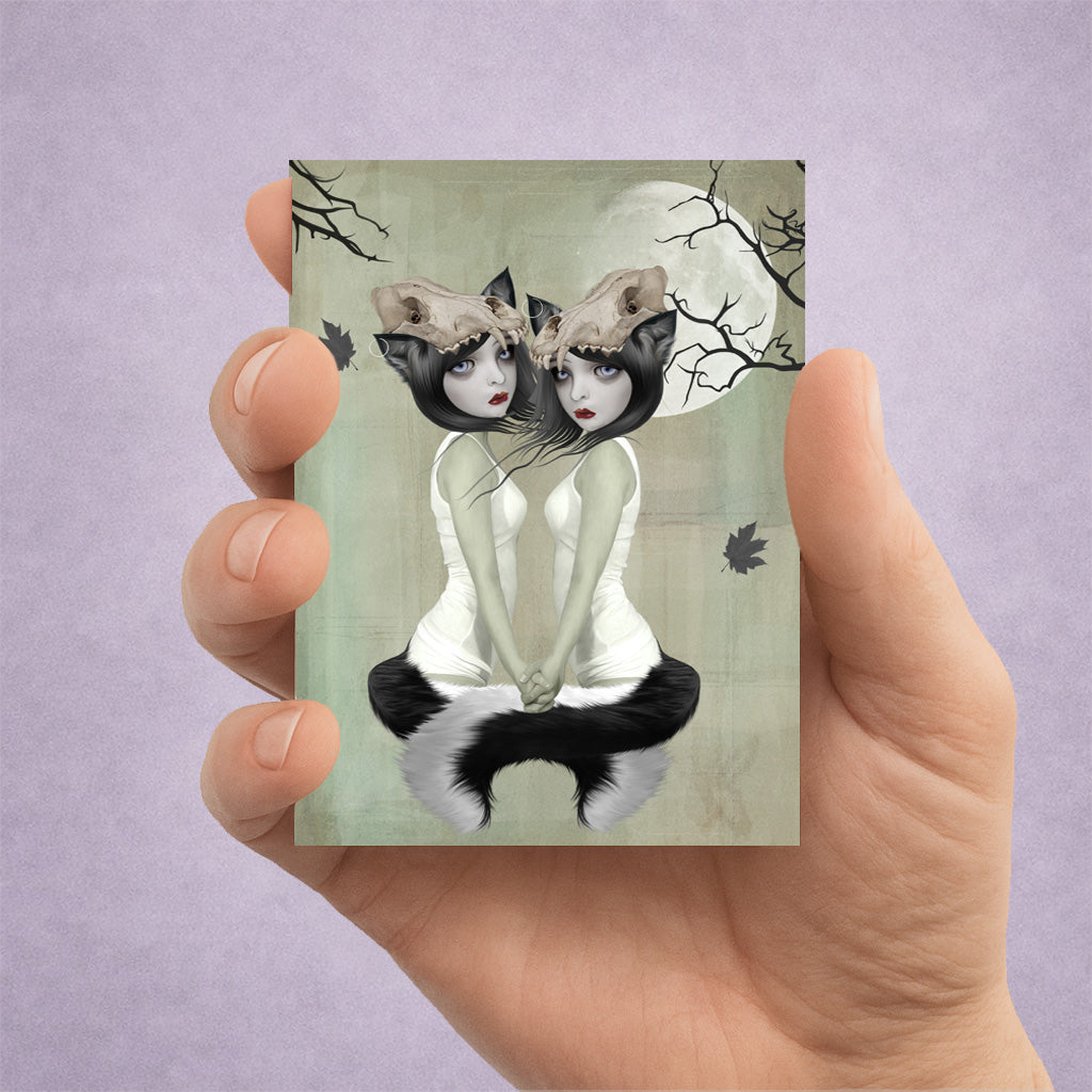 Hand holding an ACEO art card with a design of a twin werewolf sisters and a full moon on a light purple background