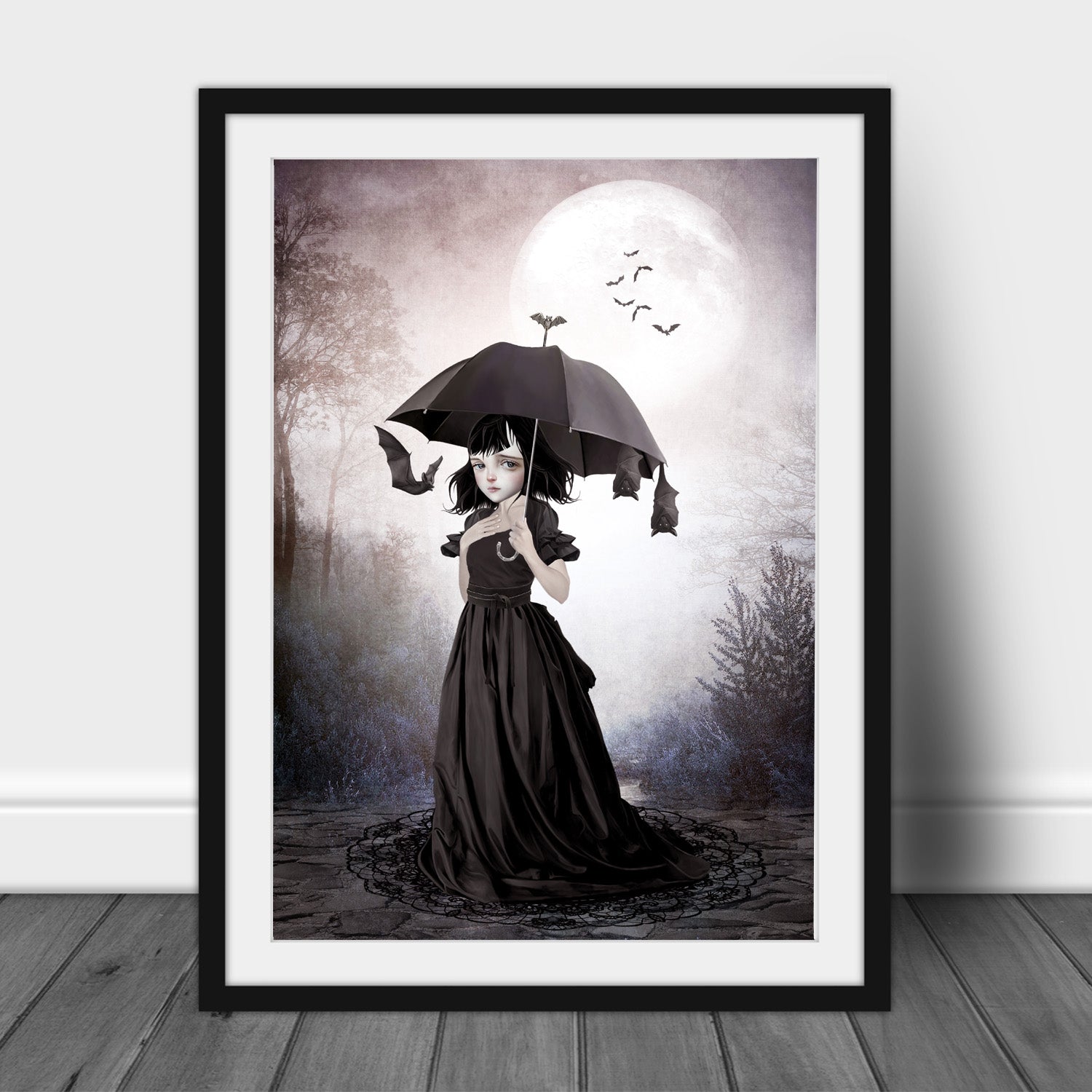 A dark fantasy art print featuring a gothic girl with a black umbrella and flying bats in a frame leaned n a white wall.