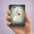 Hand holding a card with an illustration of a girl with a light bulb head surrounded by Moths displayed on  a purple background.