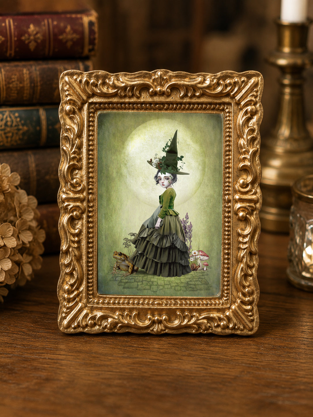 Miniature 1:12 scale cottagecore witch art print in an ornate gold frame featuring a young witch in a green dress and pointed hat, standing with a large toad and mushrooms under a full moon, displayed in a dollhouse setting.