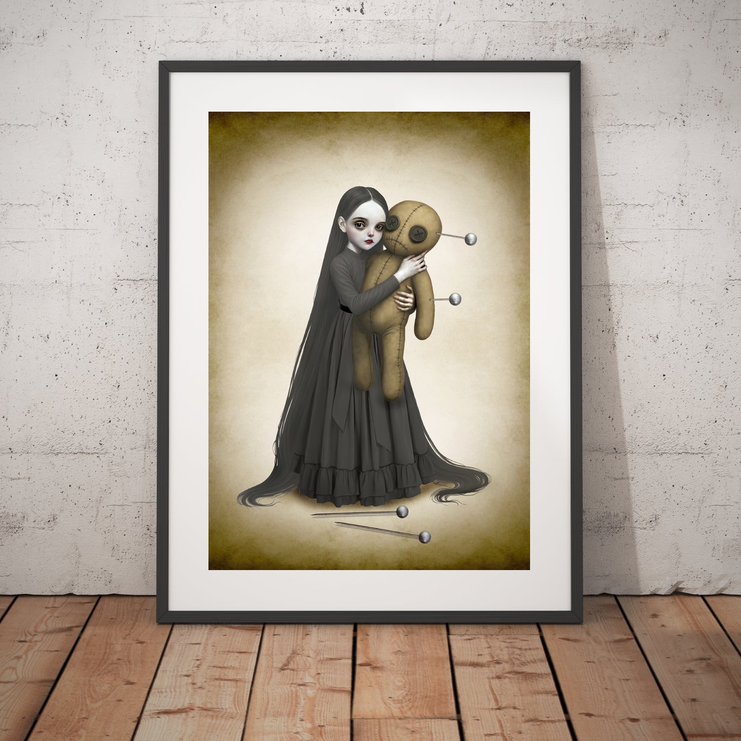 Goth girl and voodoo doll art print in black frame leaning against white wall.