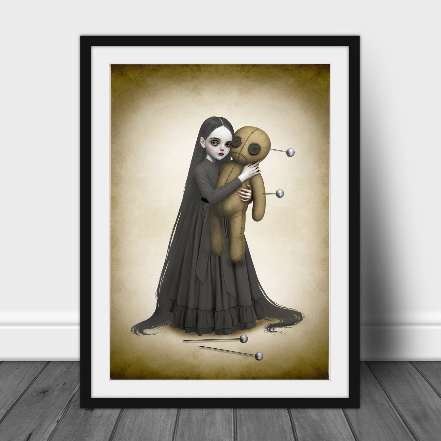 Surreal voodoo doll art print in a black frame leaning against a white wall, highlighting the eerie illustration of a long-haired gothic girl and her pinned plush doll.