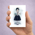 Hand holding a card with an illustration of a sailor girl holding a fishbowl with miniature shark against a light purple background 